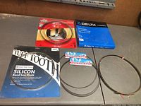 Six band saw blades displayed on table, showing original packaging for Delta Industrial, Busy Bee, and Tuff Tooth Silicone blade. Also includes unpackaged blades coiled on table.