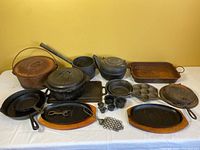 Overview of all cast iron cookware pieces grouped on white tablecloth showing pots, pans, waffle maker, muffin pan, griddles and other items