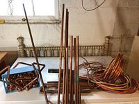Copper pipes of assorted lengths standing upright on a freezer with copper wire coils and a blue container of copper couplings visible behind.