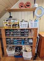 Wide shot of wooden shelving with various bins, baskets, and containers filled with craft supplies including wooden pieces, bells, glue, and hardware, plus the clamping magnifying light on the right.