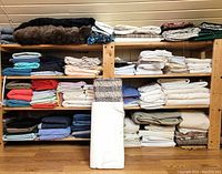 Three-tier wooden shelving unit with multiple folded fabric stacks; top shelf holds faux fur pelts and a multicolor fabric roll; middle and bottom shelves hold various fabric folds.