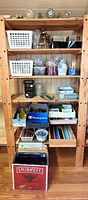 Wooden shelving unit containing baskets, boxes, tools, bottles, vases, tile strips and Smirnoff box of long glass strips