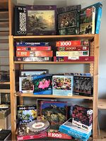 Wide view of the puzzle lot showing three wooden shelves filled with boxed jigsaw puzzles of various piece counts, brands, and themes.