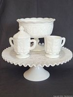 Front view of milk glass items showing the cake platter, footed candy/nut dish, creamer, and sugar holder with lid placed on the platter