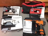 Four brad nailers and staplers arranged on a table showing different brands and manuals, including a red and black Jobmate and orange Ridgid.