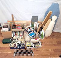 Wide shot showing assortment of sewing trims, rulers, mannequins, small irons, containers of buttons and eyelets, and various sewing tools arranged on a wooden floor.
