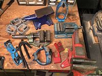 Overview of assorted vices, clamps, anvil, and clamp parts on wooden workbench showing wear and condition.