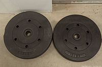 Two black weight plates side by side on concrete floor, each embossed with '15 LBS'.