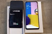 Front view of two black Android smartphones on white box, one shows Umidigi brand boot screen, the other Samsung Galaxy A21S active screen with visible crack top right corner.