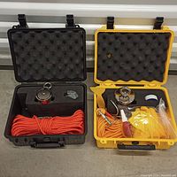 Photo shows both fishing magnet kits open side by side. The black case has a magnet, orange rope, and accessories in foam cutouts. The yellow case contains a magnet, orange rope, yellow gloves, carabiner, tape, and spray bottle.