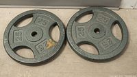 Two iron weight plates side by side on a floor, showing the 25lb markings on both plates clearly.