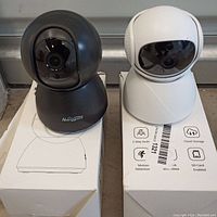 Two smart WiFi cameras, black and white, shown standing on their boxes. Cameras have dome heads and glossy front camera covers.