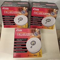 Front view of three boxes of Kidde 10-year talking smoke alarms with integrated 2-in-1 LED strobe light, showing product picture and features.