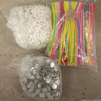 Photo showing three sealed plastic bags containing white plastic buttons, colorful pipe cleaners, and metal components on a concrete floor.