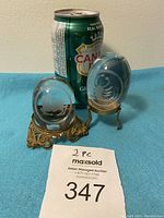 Two oval clear crystal zodiac paperweights with etched scorpion and goat designs displayed next to a soda can for size reference. Both paperweights are supported by intricate antique gold tone metal stands.
