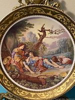 Front of porcelain plate showing detailed painted rural scene of two figures reclining near a tree by a river with ornate gold border