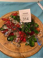 Top-down view of a wooden tray holding mixed colored chandelier crystals primarily in green, red, and clear colors with a MaxSold auction card and measuring tape visible.