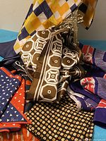Photo showing a pile of six vintage scarves featuring multiple colors and patterns including brown geometric, checkered yellow-blue, and purple with red stripes.