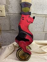 Full frontal view of the wood carving showing the bear seated on a unicycle against a concrete block wall background. The bear is painted red with black accents and a colorful purple and green top hat.