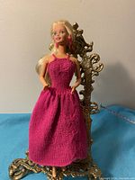 Front view of the blonde Barbie doll wearing a long pink knit dress, seated on an ornamental chair, showing detailed knit pattern on the dress and pink earrings.
