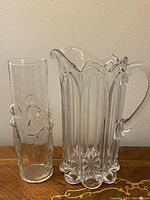 Two clear glass pieces side by side on a wooden surface: a pitcher with tulip/church window panel design, and a tall cylindrical vase with embossed dancing female figures.