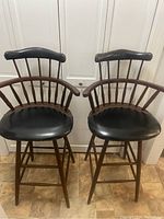 Front view of two vintage wooden bar stools side by side, showing padded black seats, top backrests with nailhead trim, wooden spindled backs and armrests.