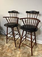 Full view of both matching vintage Duane Chair Company bar stools showing wooden frames, vertical spindles, curved arms, and black cushioned seats with brass nailhead trim.