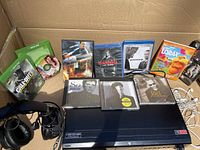 Top view of various Xbox games, movies in Blu-ray and DVD format, CDs and headphones packed in a cardboard box, featuring the black Sony blu-ray player underneath.