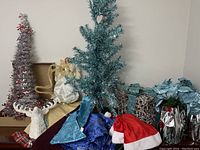 Photo showing a tall silver artificial Christmas tree with string lights, a blue-green tree, white angel tree topper, white reindeer head, blue tree skirts with snowflake pattern, red Santa hats, and wrapped silver gift box ornaments.