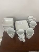 Photo showing stacked square white plates, coffee mugs, and bowls on a wood surface.
