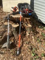 Four electric lawn tools displayed outdoors on a large tree stump and leaves. Tools include weed eater, pole saw, chainsaw, and hedge trimmer visible together.