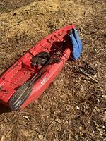 Red kayak with paddles and fishing rods attached, outdoors on dirt and leaves ground