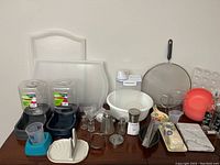 Assorted plastic pet feeders, clear plastic storage containers, white ceramic bowls, honey server, and kitchen trays on a table.