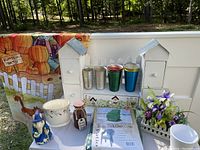 Photo showing multiple garden and outdoor decorative items including a pumpkin-themed garden flag, white wooden house-shaped shelves, metal flower holders in various colors, ceramic flower pot, resin chair covers in packaging, a blue wizard figurine, and artificial purple flowers in a white basket holder.