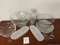 Showing all glass servingware items on wooden surface including bowls of varying sizes and patterns, pitchers, glass bottle, measuring cup, and plastic lids.