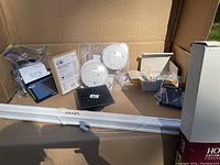 Lot of household items including two white round smoke detectors, black fan remote receiver, instruction manual, and miscellaneous hardware inside a cardboard box.