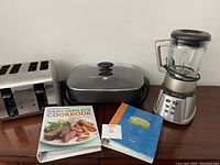 Overview of the kitchen appliances and cookbooks on a wooden surface against a light wall. Includes toaster, electric skillet, blender, and two cookbooks.