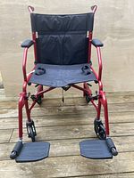 Front view of lightweight transport wheelchair showing red frame, black seat and backrest fabric, padded armrests, small caster wheels, and attached footrests.