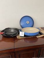 Shows all cookware items side by side on wooden cabinet: three black cast iron pans and one blue enamel coated pan with lid.