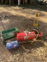 Wide shot of all four items: air tank, red gas tank with hose, yellow gas edger, green lawn spreader on leaves.