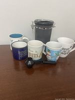 Set of five assorted coffee mugs, one black airtight coffee canister with metal clasp, and a small black coffee scoop, displayed on a wooden surface.