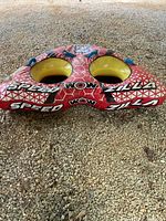 Red and yellow WOW towable tube for two, placed on gravel ground, showing front and top view