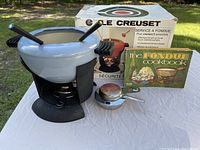 Photo showing a white enameled cast iron fondue pot on a black metal stand with burner underneath, six fondue forks, a metal burner container, a vintage fondue cookbook, and the original Le Creuset box.