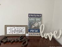 Four wooden wall art pieces arranged on a table against a wall including 'Thankful', 'Love You More', cursive 'family', and 'love' signs.