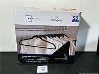 Front view of a white box with a tear on the lower right side showing 50 pack of Mainstays felt hangers with black felt hangers pictured hanging on a metal rod.
