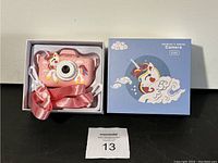 Photo of children's digital camera in original packaging showing pink camera with unicorn design, white wrist strap, and pink ribbon strap.