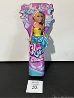 Front view of a Glimma Girlz Princess doll featuring a blonde-haired doll in yellow and blue dress attached to branded packaging with floral designs.