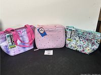 Front view of three lunch bags in different colors and patterns with tags attached, displayed on black surface against white wall.