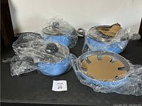 Four-piece set of light blue cookware on black surface, plastic wrapping intact to protect items.