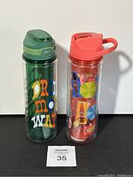 Two clear plastic water bottles side by side on a black surface against a white background. One bottle has a green lid and colorful text. The other has a coral lid and umbrella pattern.